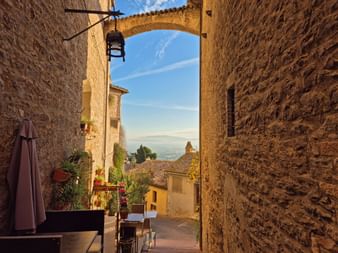 Blick durch einen Steinbogen in Assisis Altstadt mit Steinmauern, Topfpflanzen und Aussicht auf Dächer und Landschaft unter blauem Himmel.