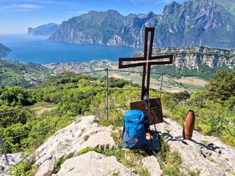 Wooden summit cross with blue backpack on rocky peak overlooking Lake Garda's northern shore, Torbole, and dramatic mountain ranges.
