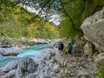 Zwei Wanderer auf felsigem Weg neben türkisfarbener Soča bei Trnovo ob Soči. Moosbewachsene Felswand rechts, üppiger Wald links.