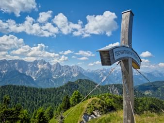 Hölzernes Gipfelkreuz am Techantinger Mittagskogel mit Panoramablick auf schneebedeckte Alpen, bewaldete Hügel und blauen Himmel mit weißen Wolken.