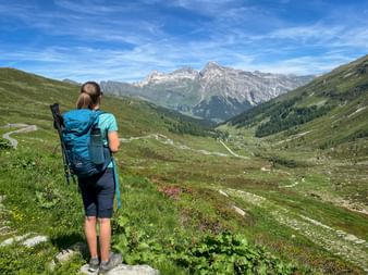 Female hiker with blue backpack and hiking poles viewing mountain valley at Splügen Pass. Green slopes lead to rocky peaks under blue sky.