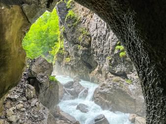 Blick durch eine Höhlenöffnung in der Partnachklamm auf tossendes weißes Wasser zwischen Felswänden mit grünem Moos und Bäumen darüber.
