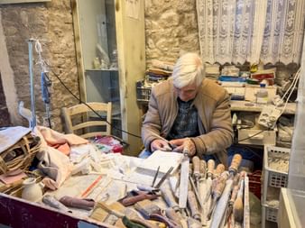 Handwerker mit weißen Haaren bearbeitet Alabaster in einer traditionellen Werkstatt in Volterra, umgeben von Schnitzwerkzeugen und Steinmaterialien.