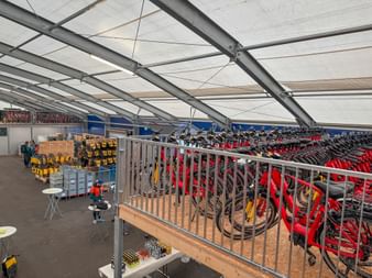 Large tent facility with rows of red bicycles stored on racks. Metal railings separate different sections, with repair equipment visible below.