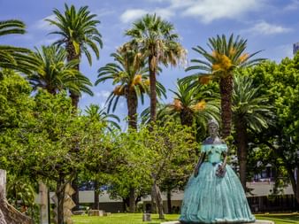 Bronze statue of Empress Sissi in turquoise ball gown standing in a tropical garden with palm trees and green lawn under blue sky in Madeira.