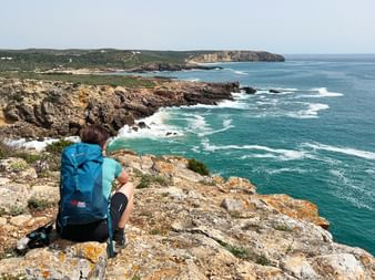 Wanderer mit blauem Rucksack sitzt auf felsiger Klippe mit Blick auf türkisfarbenen Atlantik bei Praia da Ingrina, Algarve-Küste mit Klippen.