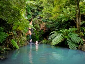 Zwei Personen baden in einem türkisfarbenen Thermalbecken in Caldeira Velha, Azoren, umgeben von üppigen grünen Farnen und tropischer Vegetation.