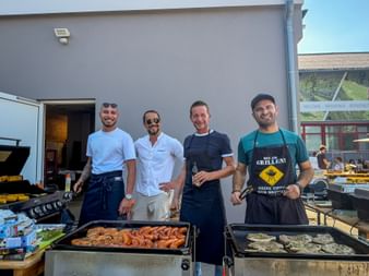 Vier Männer stehen hinter Grills mit Würstchen und anderen Speisen bei einem Sommerfest. Sie lächeln vor einem Gebäude in die Kamera.