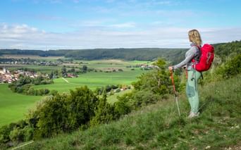 Wanderin mit rotem Rucksack und Wanderstöcken blickt auf die Altmühltal-Landschaft mit grünen Feldern, Wäldern und einem Dorf.