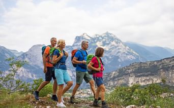 Vier Wanderer mit Rucksäcken auf einem Bergweg mit dramatischen Alpengipfeln im Hintergrund nahe dem Gardasee, Italien.