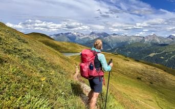 Hiker with red backpack walking on mountain trail near Obernberg with panoramic view of Alpine peaks and green slopes under blue sky.