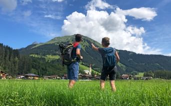 Zwei Wanderer mit Rucksäcken stehen auf einer grünen Wiese am Lechweg und zeigen auf einen Berg. Alpendorf und Kirche unter Bergen sichtbar.