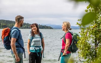 Drei Wanderer mit Rucksäcken stehen am Seeufer, umgeben von grünem Laub. Berge und bewaldete Hügel im Hintergrund sichtbar.