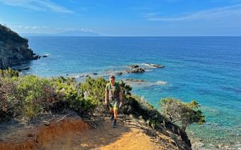 Wanderer mit Rucksack auf Küstenpfad in Cinque Terre mit türkisfarbenem Mittelmeer, felsigen Klippen und grüner Vegetation.