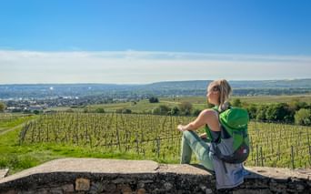 Frau mit grünem Rucksack sitzt auf Steinmauer mit Blick auf Weinberge bei Oestrich-Winkel, mit sanften Hügeln und Stadt in der Ferne.