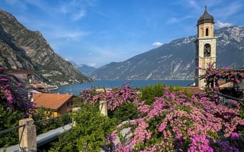 Church tower with pink flowers in Limone sul Garda overlooking Lake Garda, surrounded by mountains under blue sky.
