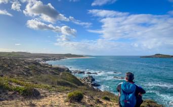 Wanderer mit blauem Rucksack sitzt auf Küstenklippe mit Blick auf türkisfarbenen Atlantik am Praia do Sissal, mit Sandstrand und felsiger Küste.