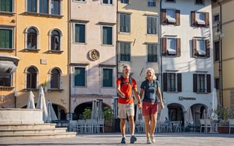Zwei Wanderer gehen Hand in Hand über einen historischen Platz in Udine, Friaul. Bunte italienische Gebäude mit grünen Fensterläden säumen die Piazza.
