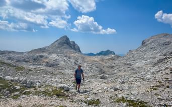 Hiker walking on rocky terrain in the Julian Alps, Slovenia. Gray limestone peaks rise under blue sky with white clouds.