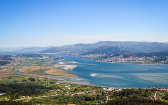 Luftaufnahme der Minho-Flussmündung mit blauem Wasser, Sandbänken, grünen Feldern und Küstendörfern. Berge erheben sich im Hintergrund.