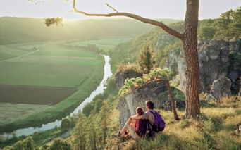 Two hikers with backpacks sitting on a cliff edge overlooking the Altmühltal valley with winding river, green fields, and forested hills at sunset.