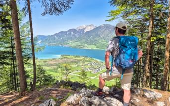 Hiker with blue backpack standing on rocky viewpoint overlooking Wolfgangsee lake in Salzkammergut, surrounded by forest and mountains.