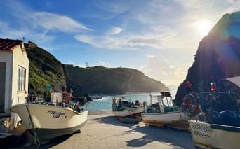 Fischerboote auf Betonpier in Porto das Barcas mit türkisfarbenem Wasser, Felsklippen und weißem Gebäude. Sonnenuntergang erzeugt Linseneffekt über der Bucht.