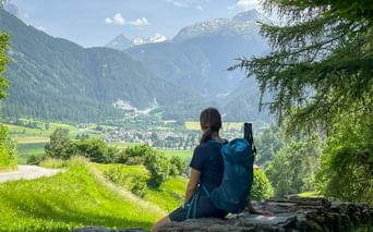 Female hiker with blue backpack sitting on rock ledge overlooking Alpine valley with village, green meadows, and snow-capped mountains.