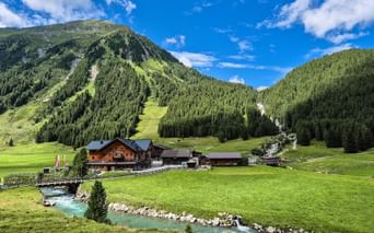 Almhütten des Krimmler Tauernhauses in grünem Tal mit türkisem Bach, umgeben von bewaldeten Bergen unter blauem Himmel in den Hohen Tauern.