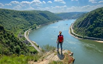 Hiker with red backpack standing on cliff edge overlooking Rhine River between Mainz and Koblenz, with forested hills and boats on the water.