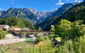Steinbrücke über türkisfarbenen Fluss im Tal der Julischen Alpen mit traditionellen Gebäuden und dramatischen Berggipfeln unter blauem Himmel.