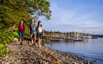 Drei Wanderer mit Rucksäcken gehen am Kieselstrand des Starnberger Sees entlang. Im Hintergrund sind ein Hafen mit Segelbooten und Bäume sichtbar.