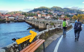 Zwei Wanderer mit Rucksäcken auf der Brücke Ponte Sampaio mit gelbem Camino Peregrino-Schild. Steinbrücke überquert Fluss mit Hügelstadt im Hintergrund.