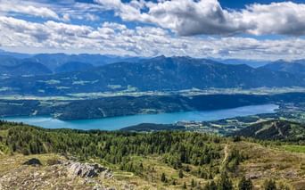 Panoramic view from Gerlitzen mountain showing a large blue lake surrounded by forested hills and distant Alpine peaks under cloudy sky.