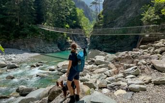 Female hiker with dog and blue backpack standing on rocks at Viamala Gorge, viewing suspension bridge over turquoise Hinterrhein river.