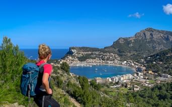 Female hiker with blue backpack viewing Port de Sóller bay from hillside on GR221 trail in Mallorca. Mountains and Mediterranean Sea visible.