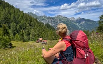 Female hiker with burgundy backpack sitting in alpine meadow near Trins, Gschnitztal, viewing mountain peaks and traditional huts below.