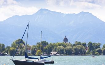 Blaues Segelboot auf dem Chiemsee mit Kirchenkuppel auf bewaldeter Insel und Bergkette im Hintergrund unter teilweise bewölktem Himmel.
