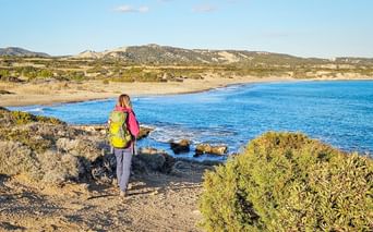 Wanderin mit gelbem Rucksack auf Küstenweg mit Blick auf blaue Bucht mit Sandstrand und Hügeln in Zypern.