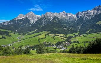 Panoramic view of Maria Alm valley with green meadows, scattered villages, and dramatic rocky mountain peaks under blue sky.