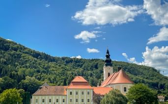 Engelszell Abbey complex with yellow monastery building, church with orange roof and black onion dome tower, set against forested hills.