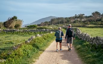 Two hikers walking on a gravel path between stone walls near Oia. Green fields and mountains are visible in the background under a cloudy sky.