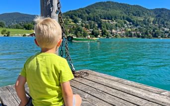 Blondes Kind im gelben Shirt sitzt auf Holzsteg am Tegernsee. Türkisfarbenes Wasser, grüne Berge und Dorf im Hintergrund unter blauem Himmel.