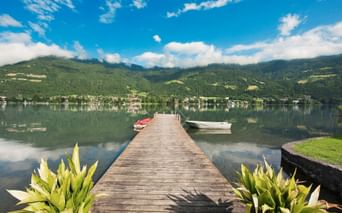 Wooden pier extending into calm Wörthersee lake with boats moored nearby. Green mountains and village reflected in water under blue sky.