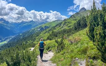 Female hiker with backpack walking on mountain trail through green alpine landscape with snow-capped peaks and blue sky with white clouds.