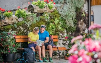 Two hikers sitting on a wooden bench studying a map, surrounded by colorful flowers and plants on a stone wall in the Alpe-Adria region.