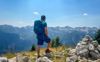 Hiker with backpack standing on rocky summit in the Julian Alps, overlooking mountain ranges under blue sky with stone cairn nearby.