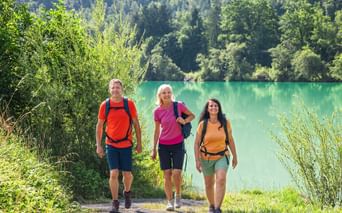 Three hikers with backpacks walking on a path along the turquoise Danube River. Dense green forest covers the hillside in the background.