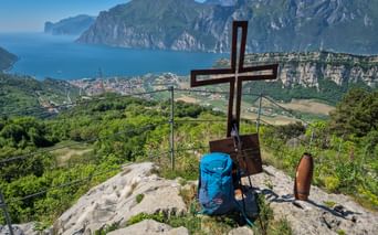 Wooden summit cross with blue backpack on rocky peak above Lake Garda. View shows Torbole-Nago, mountains, and northern lakeshore.