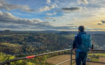 Wanderer mit blauem Rucksack auf der Lilienstein Aussichtsplattform mit Blick auf bewaldete Täler und Hügel unter dramatischem Wolkenhimmel.